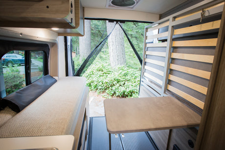 Cozy RV rental interior showing a bed, table, and open screened door looking out to trees and lush greenery.