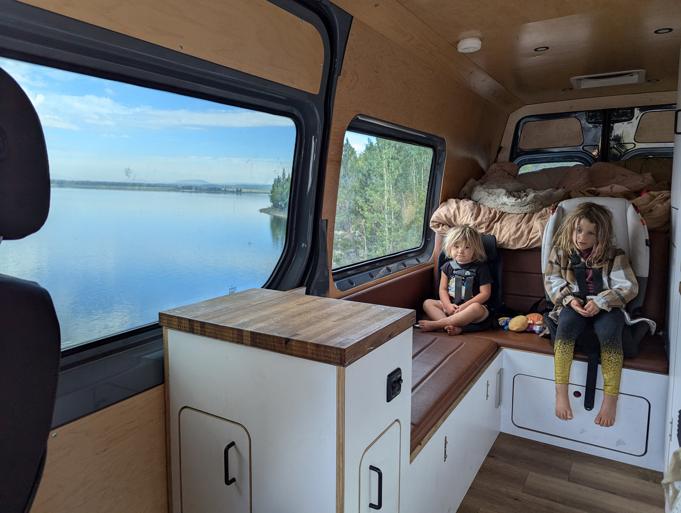 Two children relax in a spacious RV rental, enjoying scenic lake and forest views from inside their camper van. — photo 17