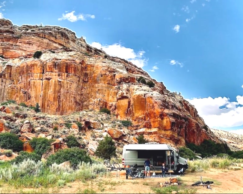 RV rental adventure! A camper van is parked at the base of a massive red rock cliff, with people and bikes outside. — photo 1