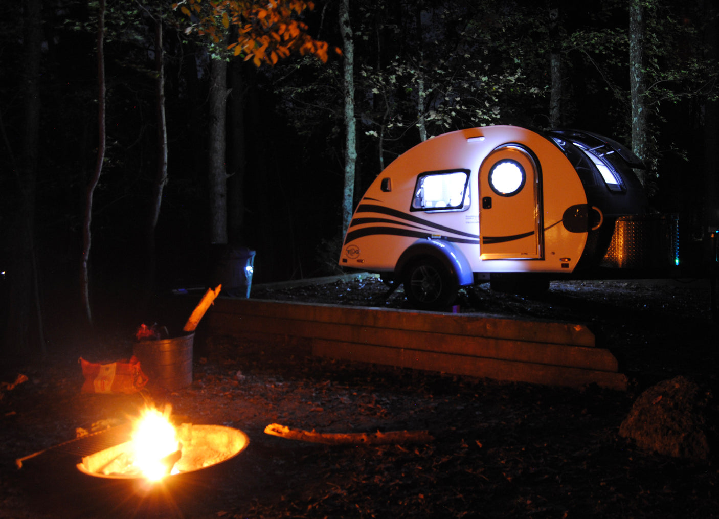 Enjoying an RV rental adventure: a lit teardrop camper parked in the woods, with a warm campfire glowing nearby.