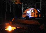 Enjoying an RV rental adventure: a lit teardrop camper parked in the woods, with a warm campfire glowing nearby.