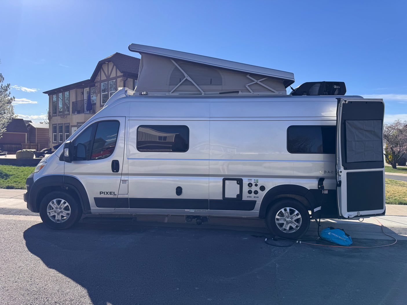 Silver camper van, potentially an RV rental, with a pop-top roof and open rear doors, connected to a fresh water source.