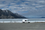 White RV rental van by a partially frozen lake, framed by snow-capped mountains under cloudy skies.