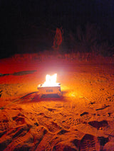 Night camping with an RV rental: a portable fire pit illuminates sandy ground with footprints and tracks, surrounded by dark foliage.