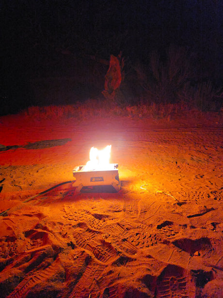 Night camping with an RV rental: a portable fire pit illuminates sandy ground with footprints and tracks, surrounded by dark foliage.