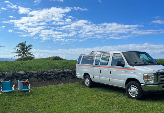 Enjoy a beach getaway! White RV rental van parked on green grass by the ocean, with chairs & palm tree.