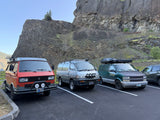 Diverse camper vans, including possible RV rental options, parked at the base of a towering rock face in a scenic lot.