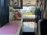 Inside an rv rental: a cozy bed at the rear overlooks a lake, with a kitchen counter & sink on the left. Ideal for travel.