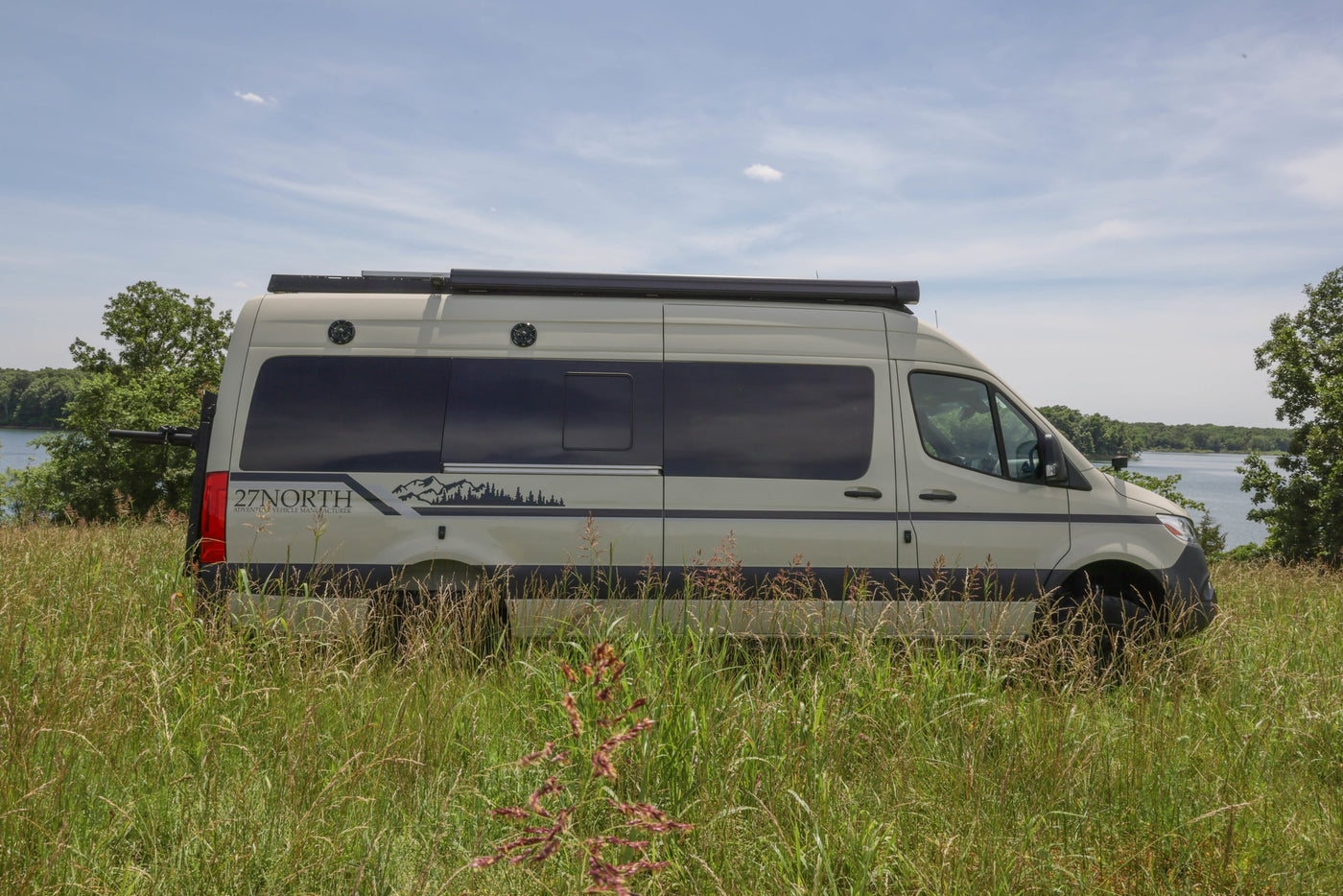 A light-colored RV rental camper van, branded "27NORTH", sits in tall grass beside a lake, ideal for outdoor adventures. — photo 1