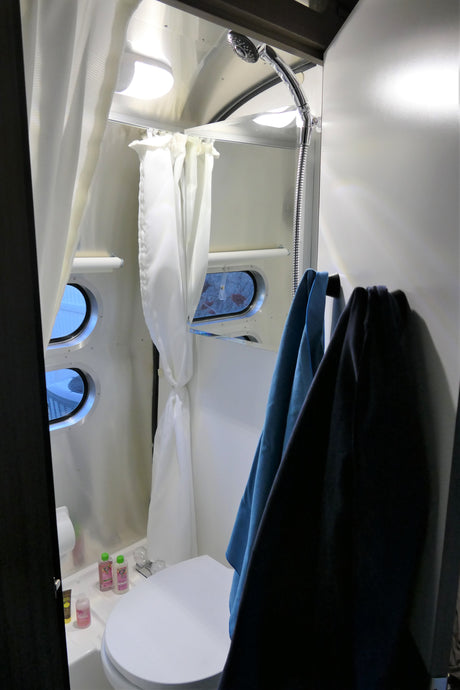This compact RV rental wet bath features a toilet, shower, privacy curtains, porthole windows, and hanging towels.