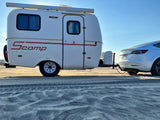 RV rental: A white Scamp travel trailer towed by a white Tesla on a sandy beach. Ideal for your next eco-camping trip!