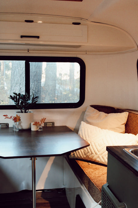 Cozy RV rental interior: dark wood table, bench seating with pillows, and window view of trees. White cabinets & small sink.