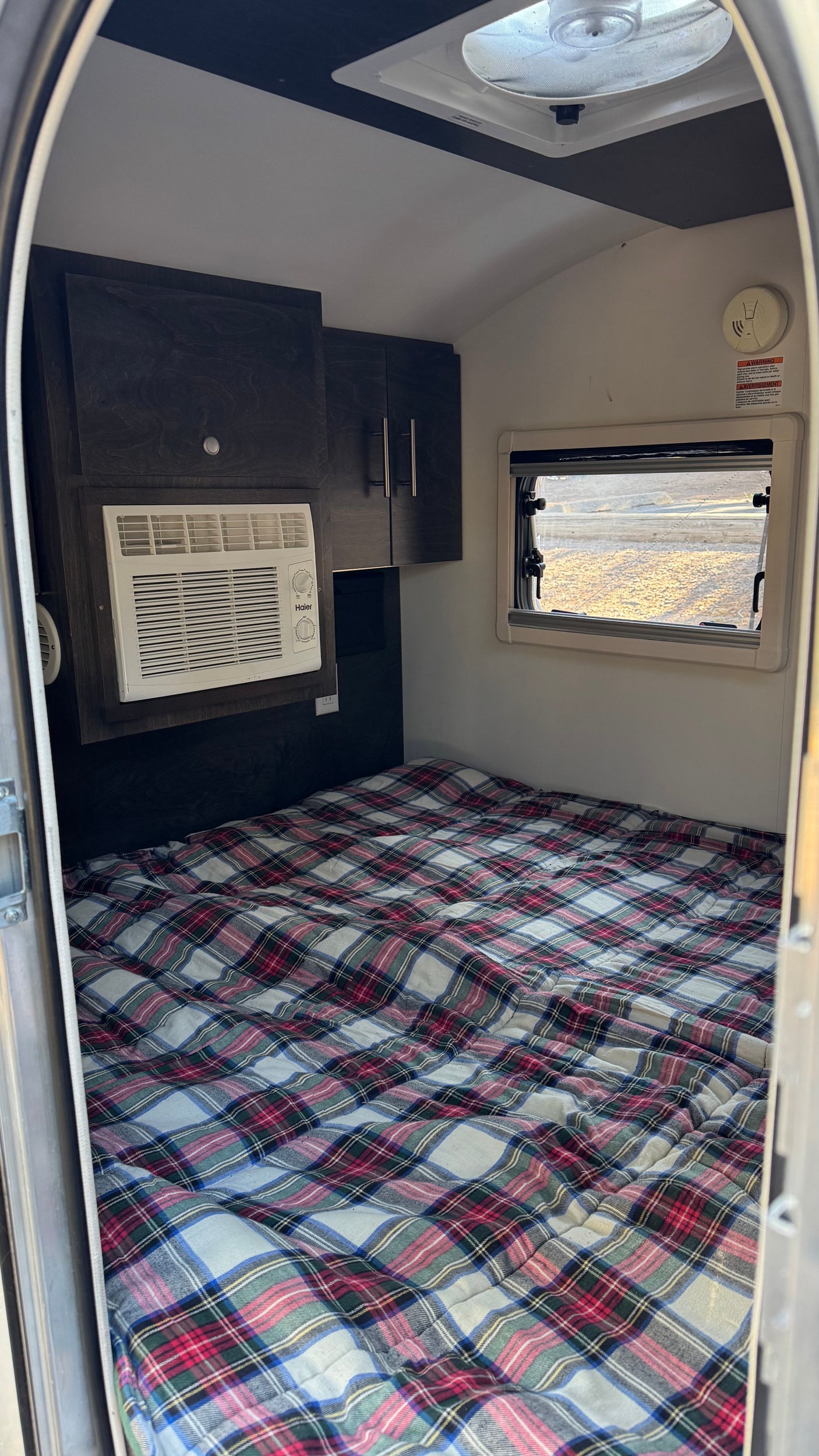 RV rental sleeping area: plaid bed, Haier AC, dark cabinets, window, & ceiling fan. Compact camper comfort.