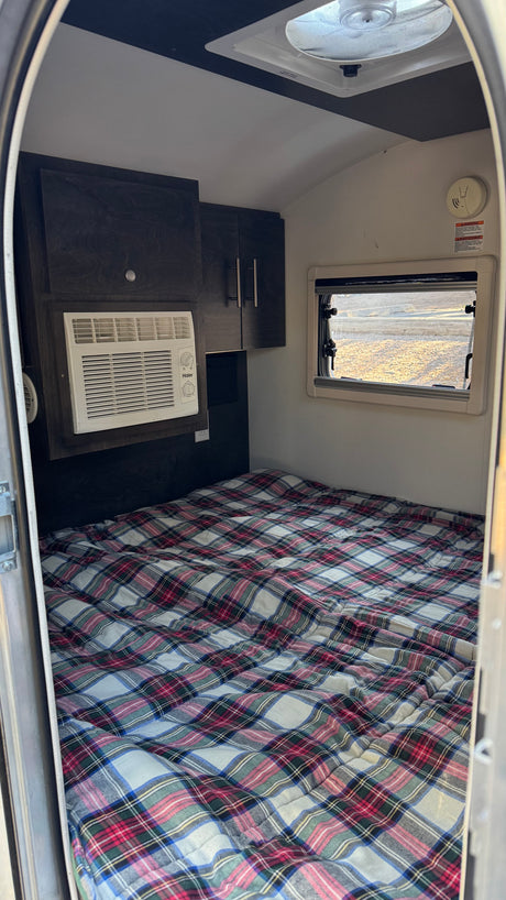 RV rental sleeping area: plaid bed, Haier AC, dark cabinets, window, & ceiling fan. Compact camper comfort.