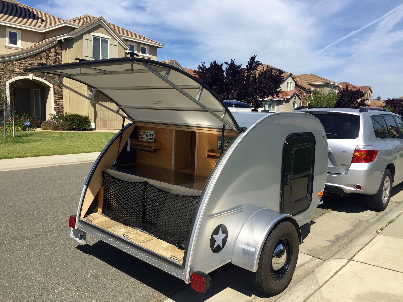 Silver teardrop camper with open galley, a unique RV rental, parked on a sunny street next to an SUV.