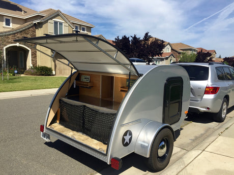 Silver teardrop camper with open galley, a unique RV rental, parked on a sunny street next to an SUV.