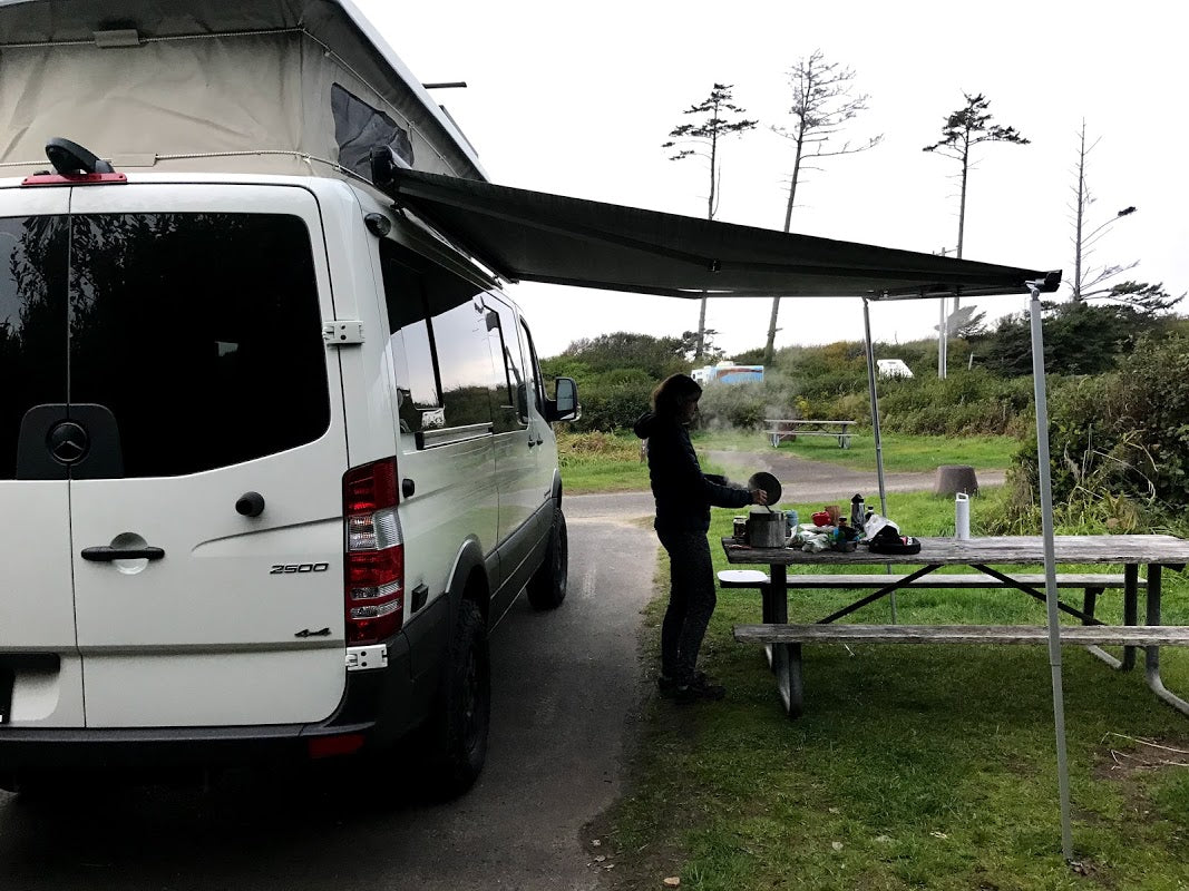 White Sprinter camper van, possibly an RV rental, with an extended awning. A person cooks on a picnic table at a campsite.