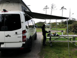 White Sprinter camper van, possibly an RV rental, with an extended awning. A person cooks on a picnic table at a campsite.