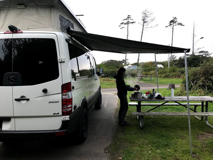 White Sprinter camper van, possibly an RV rental, with an extended awning. A person cooks on a picnic table at a campsite.