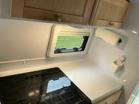 Compact rv rental kitchen features a stovetop, window with blinds, wooden cabinets, and paper towel dispenser.