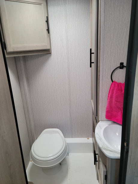 Compact RV rental bathroom with toilet, sink, and overhead storage. Features light grey walls and a bright pink towel.
