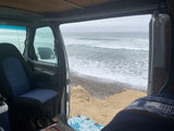 Experience beach camping with an RV rental! Van interior shows a comfy seat with waves crashing on the rocky shore outside.