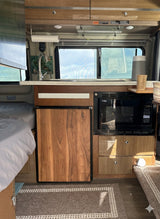 Cozy RV rental interior: kitchenette with sink, microwave, wood-paneled fridge, overhead cabinets, and adjacent bed.