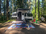 Scenic RV rental camping spot among tall trees with a picnic table, grill, and a furry friend enjoying the outdoors.