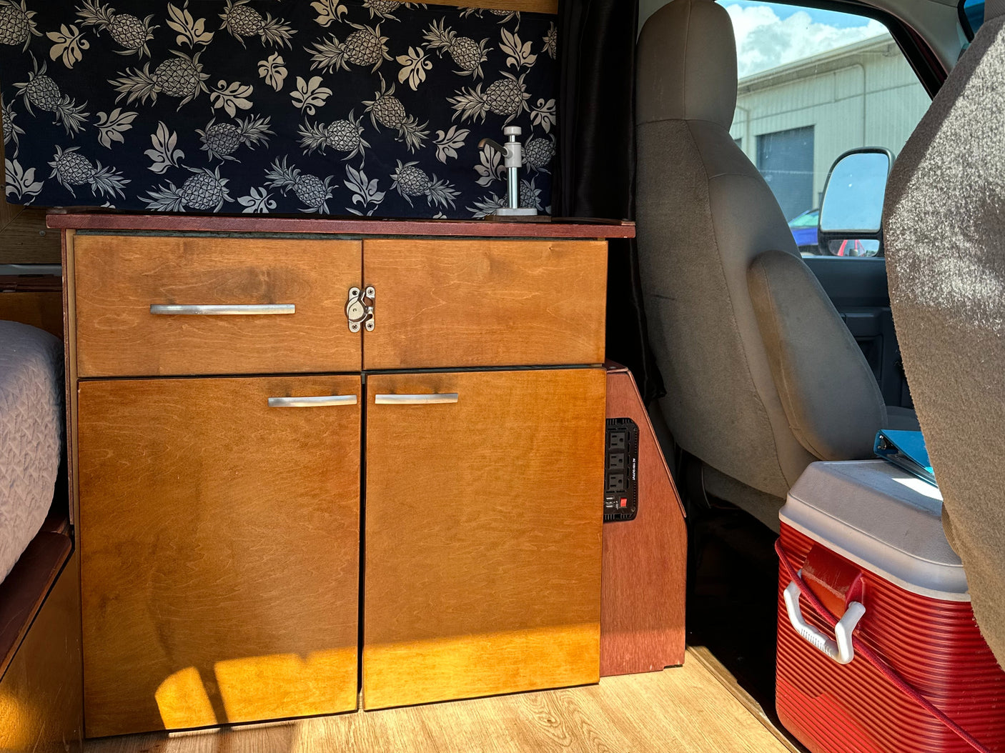 An RV rental van interior featuring custom wooden cabinets, pineapple-patterned fabric, a power inverter, and a red cooler.