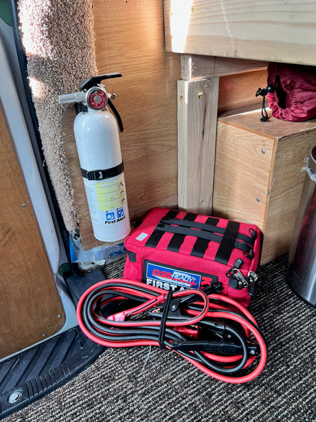 RV rental interior shows essential safety gear: a white mounted fire extinguisher, red first aid kit, and jumper cables on the floor.