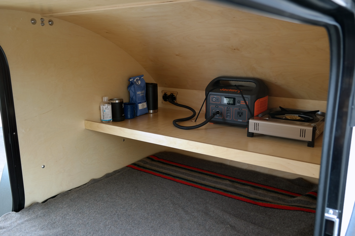 Inside a cozy RV rental: a shelf with a Jackery power station, portable stove, coffee, and bedding below.