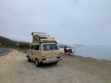 Enjoy a coastal view from your RV rental. Tan VW Westfalia parked by ocean; person in chair watches misty coast.