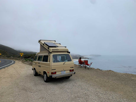Enjoy a coastal view from your RV rental. Tan VW Westfalia parked by ocean; person in chair watches misty coast.
