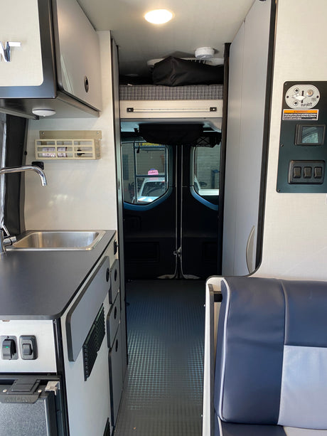 Interior of an RV rental: Compact kitchenette with sink on the left, an adjustable bed/loft above, and front cab view.