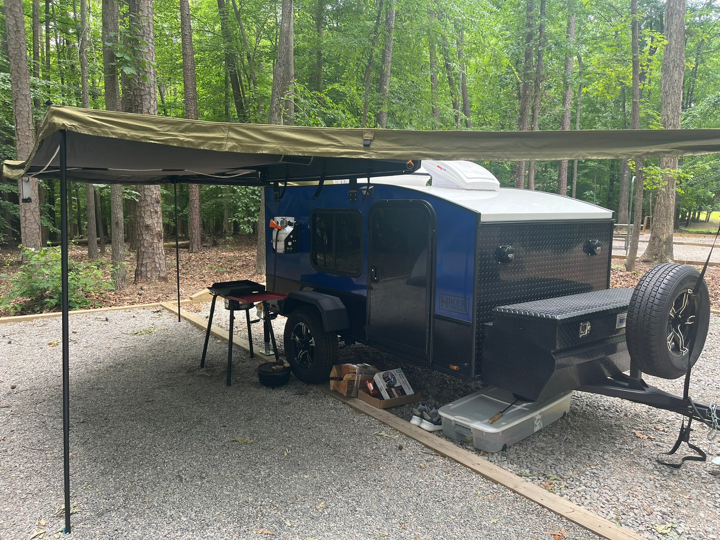 Enjoying the outdoors with this blue Hiker Teardrop Trailer RV rental at a wooded campsite, complete with an extended awning and cooking setup.