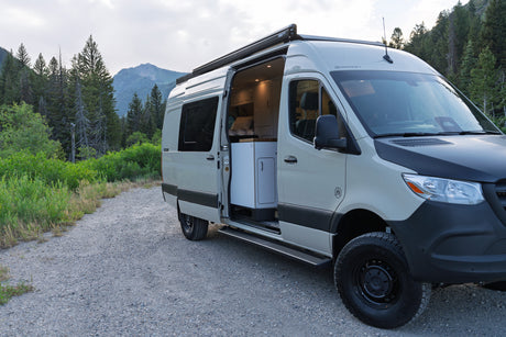 Adventure awaits! This custom camper van, perfect for rv rental, is parked on a gravel road amidst a beautiful mountain forest.