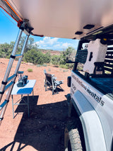 An **overland RV rental** setup: white truck with a rooftop tent, ladder, and outdoor kitchen in a sunny, arid landscape.