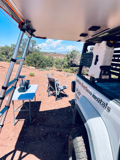 An **overland RV rental** setup: white truck with a rooftop tent, ladder, and outdoor kitchen in a sunny, arid landscape.