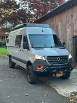 Custom Mercedes Sprinter van on an asphalt driveway. This adventure-ready rv rental features off-road tires, a winch, and a roof rack.