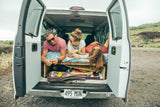 Three friends smile, relaxing in the open back of their Hawaii rv rental van. A perfect van life road trip with gear.