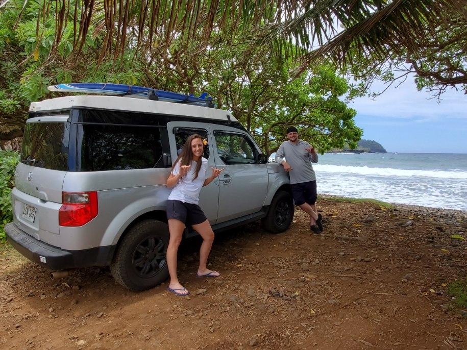 Couple with surfboards by their RV rental, a Honda Element, enjoying a scenic stop at a lush Hawaiian beach. Perfect for adventure!