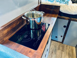 RV rental kitchen: compact setup with induction cooktop, pot on wooden counter, and adjacent sleeping area.