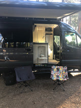 RV rental camper van with open side, revealing a lit interior kitchen. An awning extends over outdoor camping chairs.