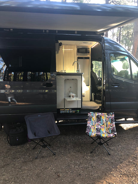 RV rental camper van with open side, revealing a lit interior kitchen. An awning extends over outdoor camping chairs.