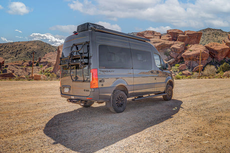 Gray Mercedes-Benz Sprinter rv rental with bike rack parked on gravel road. Red rock mountains in sunny desert.