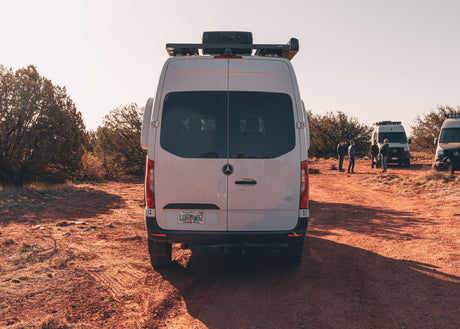 Rear view of a white Sprinter RV rental on a red dirt road, with a group and other vans in a sunny desert setting.