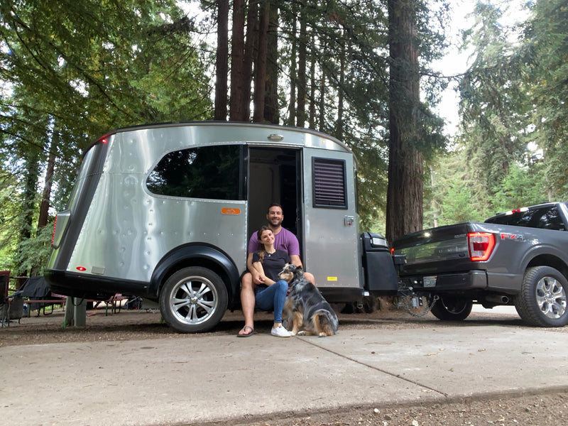 A happy couple and their dog pose by their modern silver rv rental, hitched to a truck in a scenic wooded campsite. — photo 1