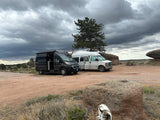 Two camper vans, one an RV rental, parked on a dirt road in a rocky landscape under dark clouds. A dog is visible in the foreground.