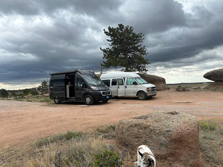 Two camper vans, one an RV rental, parked on a dirt road in a rocky landscape under dark clouds. A dog is visible in the foreground.