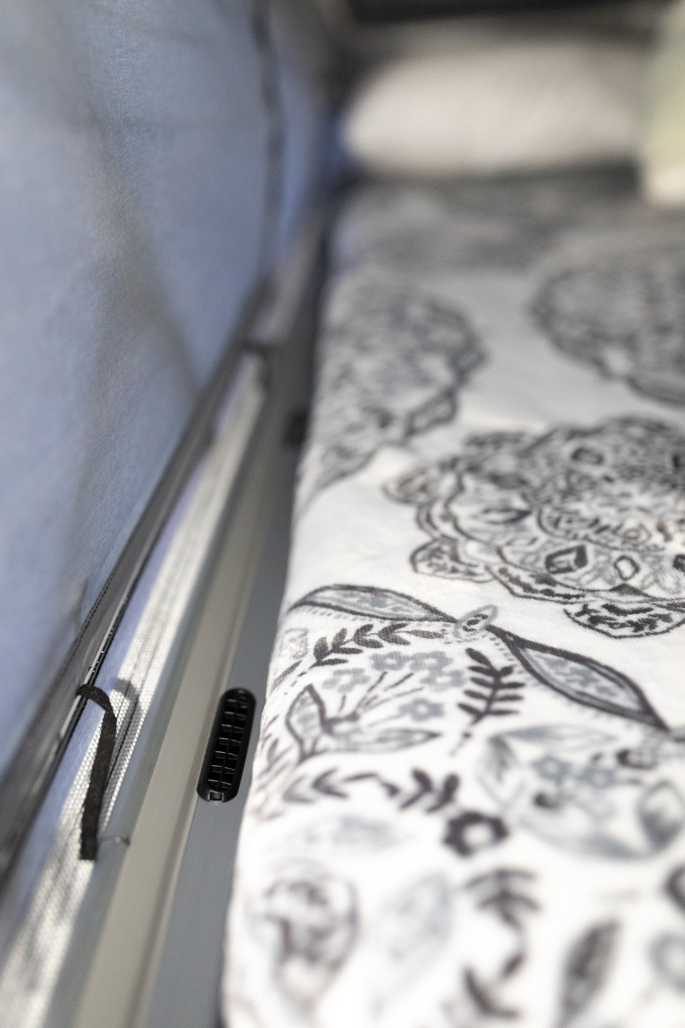 Inside an RV rental: A patterned bed with white linens borders a light wall, featuring a vent and a mesh screen.